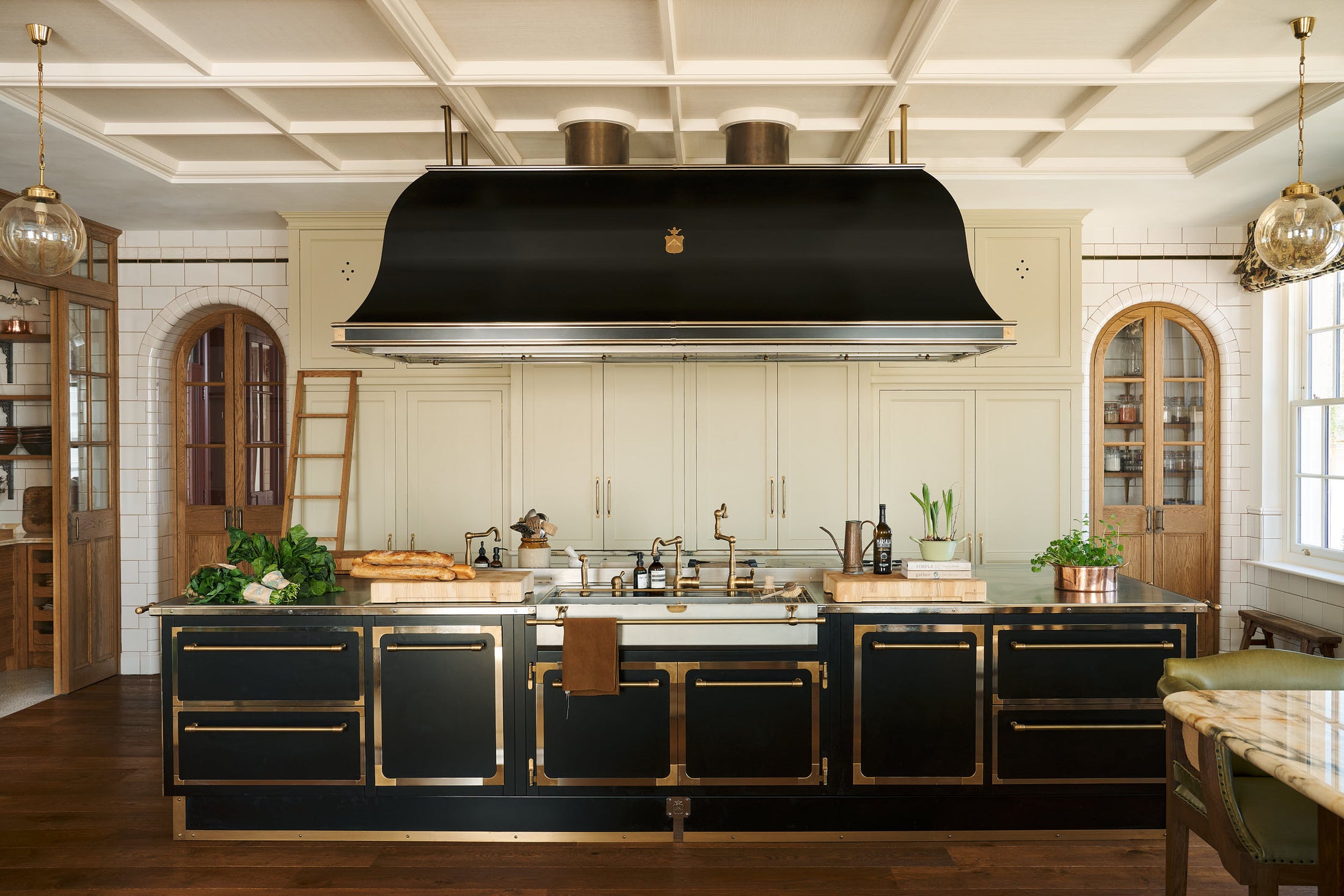 A grand room with high panelled ceiling, traditional glossy white tiled walls with curved archways on either side of a huge bank of creamy mushroom fitted full-height Classic cupboards with a polished oak ladder. An elaborate black with gold accent kitchen island and canopy sits in the middle of the